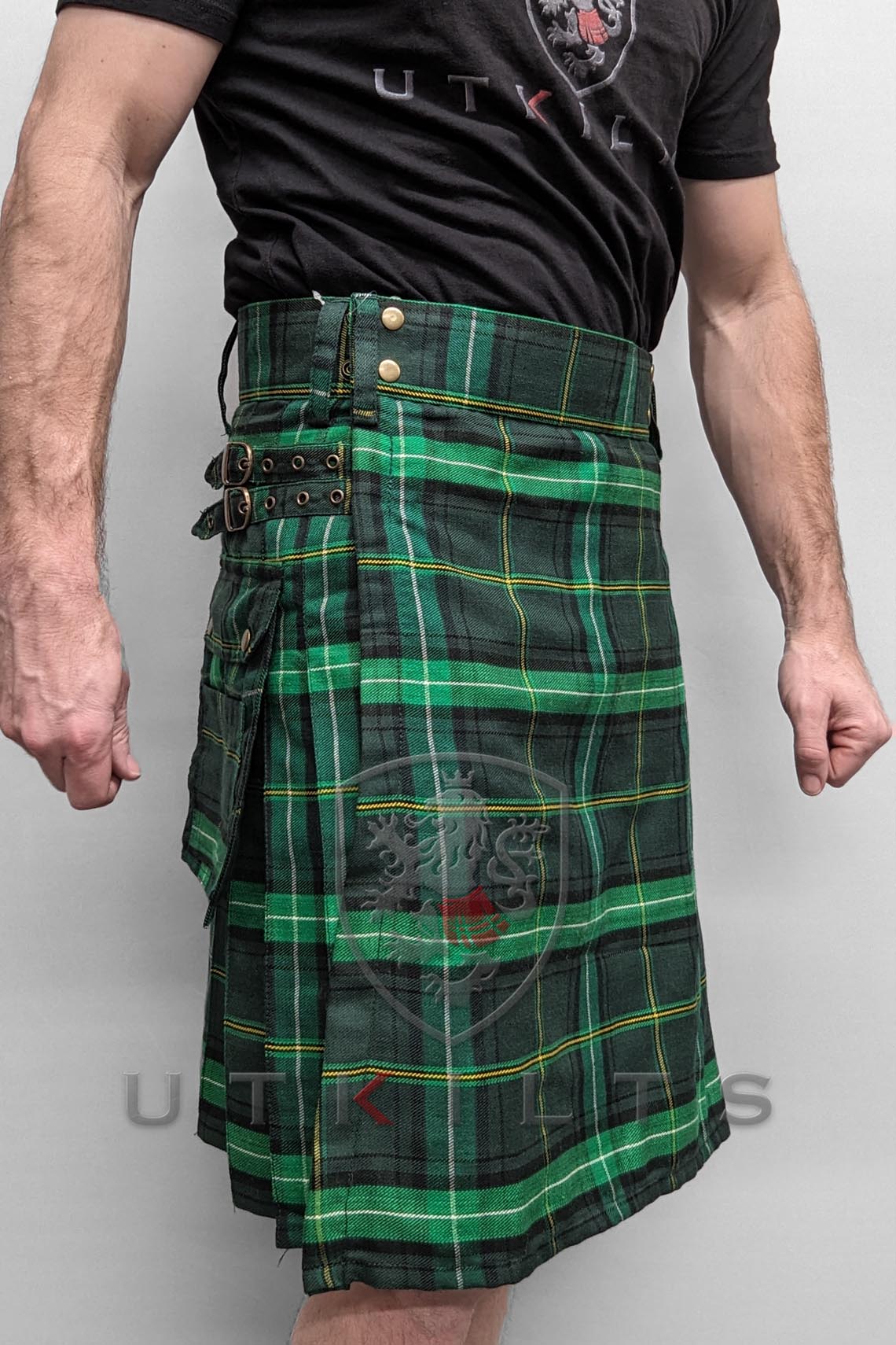 A man dressed in a green and black tartan kilt stands confidently, showcasing traditional Scottish attire.