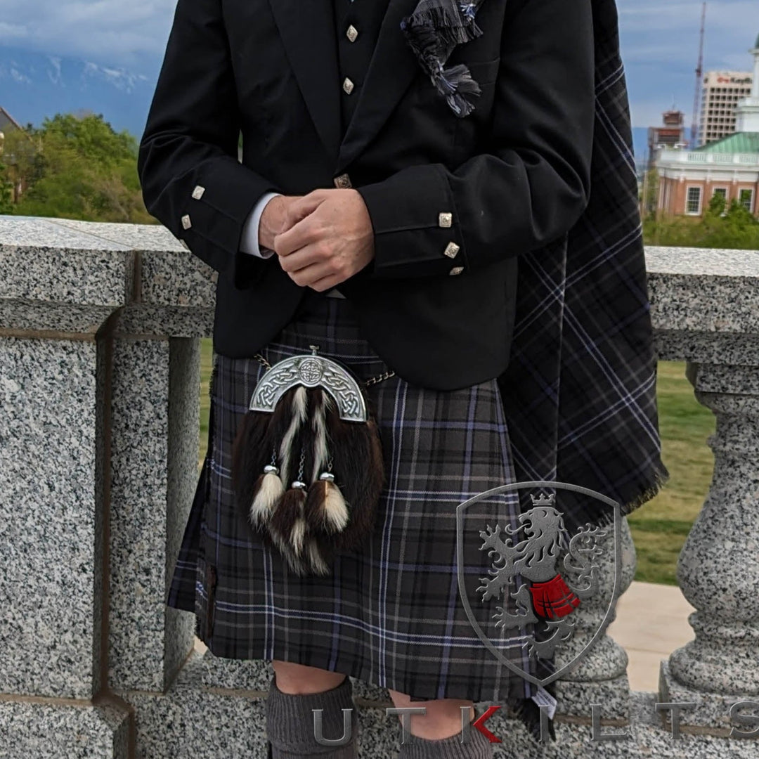 A man in a kilt stands confidently on a bridge, showcasing traditional attire against a scenic backdrop.