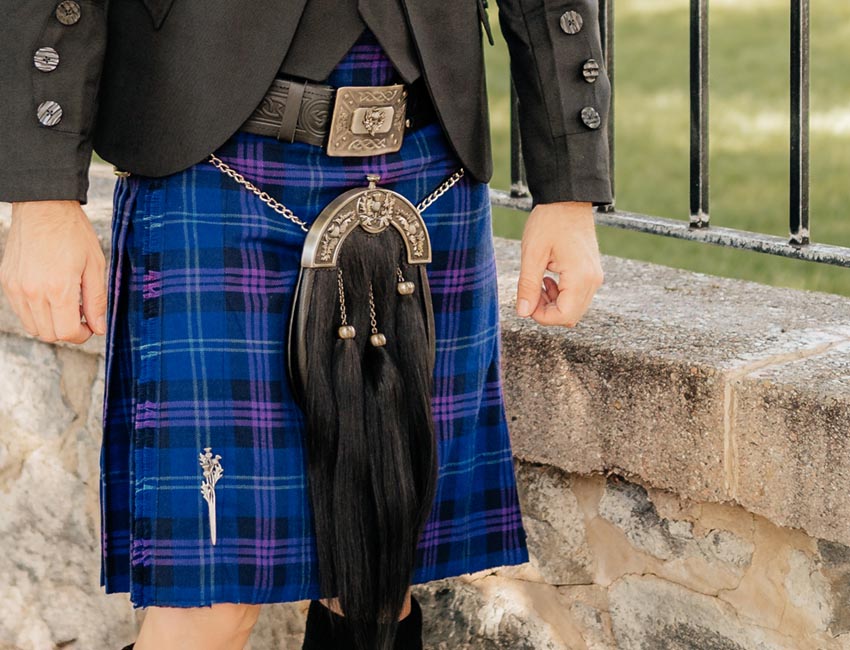 A man in a kilt stands confidently on a stone wall, showcasing traditional attire against a rugged backdrop.