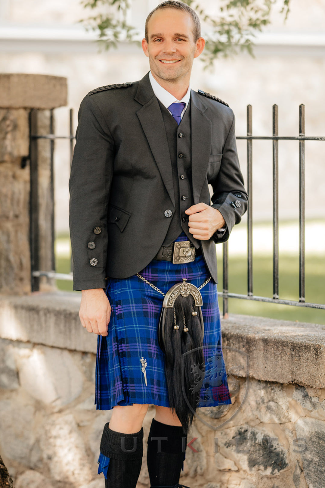 A man in a kilt stands confidently, smiling for the camera in a picturesque outdoor setting.