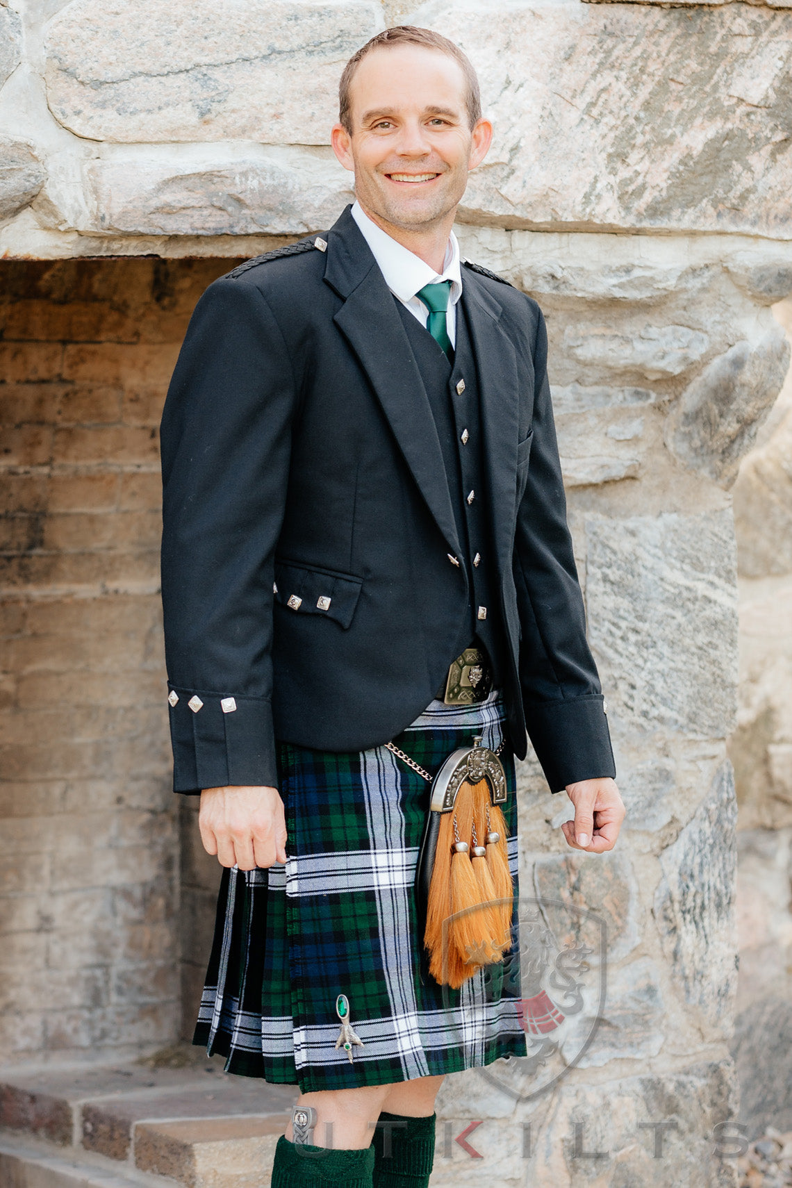 A man wearing a kilt and jacket stands confidently in front of a rustic stone wall.