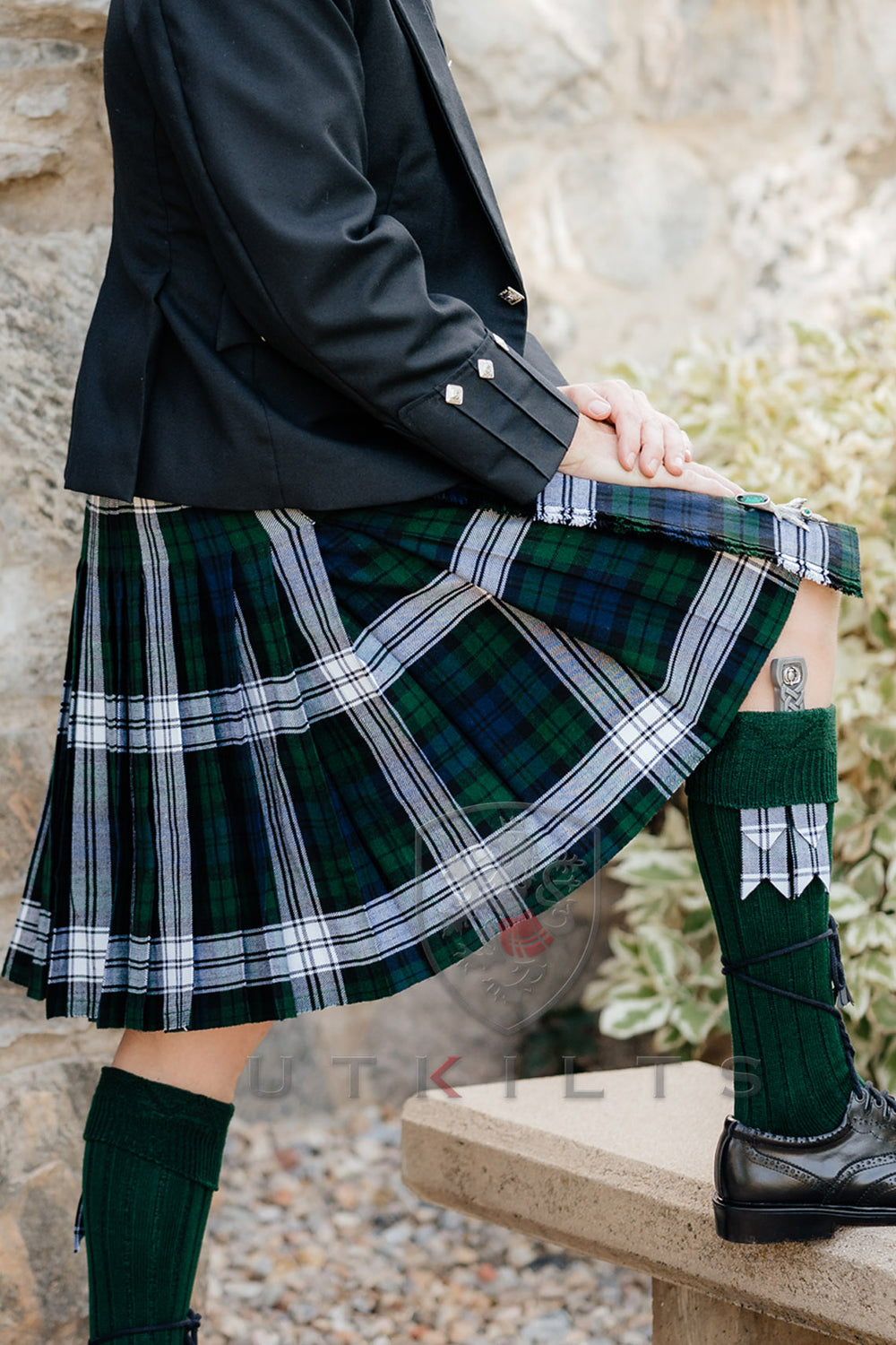 A man in a kilt and black shoes is seated on a bench, appearing calm and contemplative in a public space.