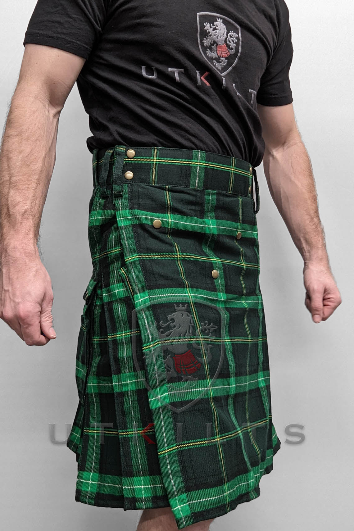 A man dressed in a green and black tartan kilt stands confidently, showcasing traditional Scottish attire.