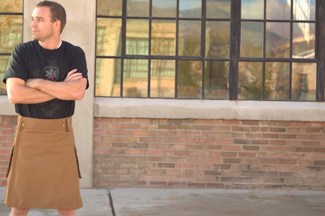 A man wearing a tan kilt poses in front of a building, highlighting the unique utility kilts available from UTKilts.
