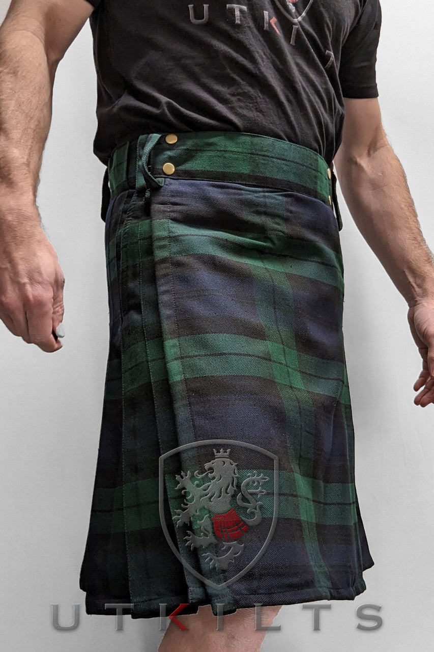 A man showcases a Business Edition kilt, featuring a black shirt and a striking green and blue tartan pattern.