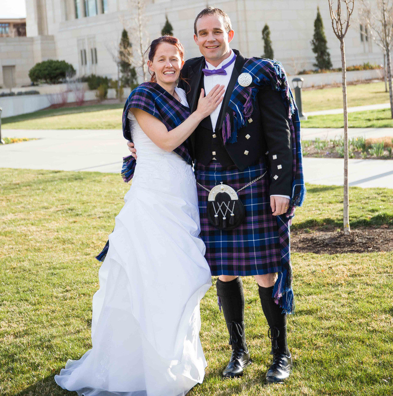 A man and woman in kilts smile for a photo, featuring special order tartan fabric, fly plaids, sashes, and neck ties.